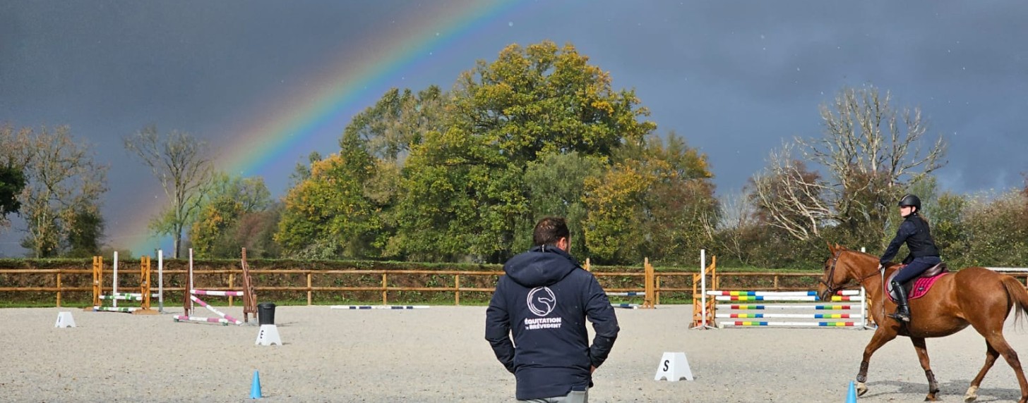 Cavalier en cours d’équitation dans la grande carrière du Brévedent sous un arc-en-ciel, encadré par un coach.